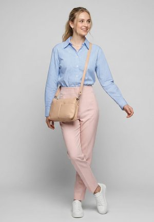 Ung kvinde i blå ternet skjorte, lyserøde bukser med høj talje, hvide sneakers, smilende og bærende en beige crossbody taske, står på grå baggrund.