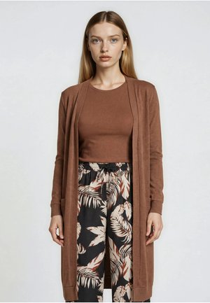 Junge Frau mit glattem blondem Haar, die einen braunen langen Cardigan, passenden Oberteil und schwarze Hose mit beige-braunen Blattmustern trägt.