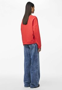 Sudadera roja con un corte relajado, mangas oversized y puños de canalé, combinada con jeans de pierna ancha en color azul descolorido. Diseño minimalista sin patrones visibles.