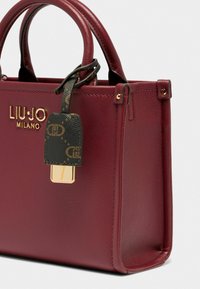 Sac à main en cuir bordeaux avec poignées courtes, détail logo en or et étiquette de bagage noire à motif. Texture lisse et forme structurée.