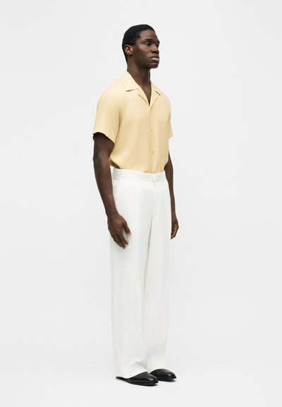 Homme debout, portant une chemise à manches courtes jaune pâle boutonnée, un pantalon blanc à jambes larges, et des chaussures noires sur un fond blanc uni.