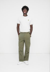 T-shirt blanc avec un petit logo jaune, pantalon cargo vert olive avec poches latérales, et baskets blanches. Tenue simple et fonctionnelle.