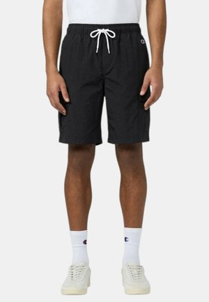 Uomo che indossa pantaloncini cargo neri con coulisse, calzini bianchi Champion, sneakers bianche e camicia bianca, in piedi davanti a uno sfondo monocromatico.