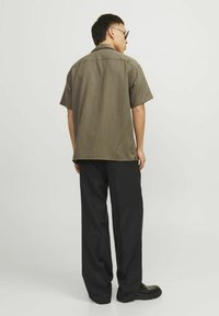 Vyras vilkintis alyvuogių spalvos trumpomis rankovėmis marškiniais, juodomis plačiomis kelnėmis, juodais batais ir akinių nuo saulės stovi atsisukęs į baltą foną.