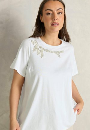 Frau mit langem braunem Haar, die ein weißes Kurzarmshirt mit silberner Perlenstickerei nahe dem Halsausschnitt trägt, steht vor einem neutralen Hintergrund.