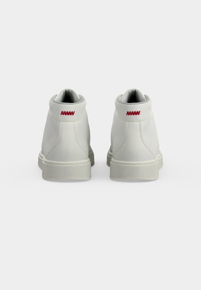 Paire de baskets montantes blanches vues de dos avec un détail de logo rouge sur la languette du talon sur un fond clair uni.