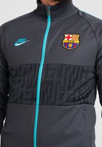 Czarna bluza Nike z zamkiem błyskawicznym w ciemnoszarym kolorze z turkusowymi akcentami, z herbem Barcelony oraz teksturowanym geometrycznym wzorem na dolnej części.