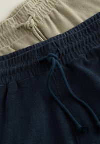 Deux paires de pantalons de survêtement avec cordon de serrage : une en vert olive clair, l'autre en bleu marine. Les deux possèdent des ceintures côtelées et des textures douces.