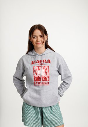Jonge vrouw met bruin haar die hoepel oorbellen draagt, een grijze hoodie met rode basketbaltekst en groen gestreepte korte broek, staand met handen in de zakken.