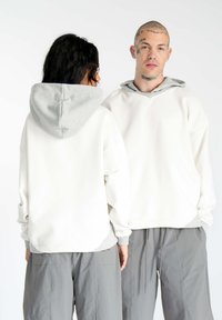 Hvid oversized hoodie med en grå indvendig hætte, der har dropped shoulders og manchetterede ærmer. Stylet med grå bukser, minimalistisk design.