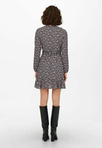 Žena s kudrnatými vlasy, oblečená v dlouhém květinovém šatech s dlouhým rukávem a černými vysokými podpatky, stojící zády.