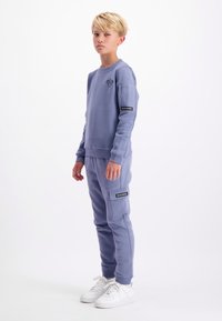 Blonde jongen die staat in een bijpassende blauwe sweatshirt en joggers met zwarte patches, gecombineerd met witte sneakers, tegen een witte achtergrond.