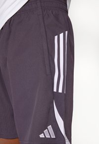 Közeli kép sötét sportnadrágról, fehér háromcsíkos mintával és oldalt hímzett Adidas logóval.