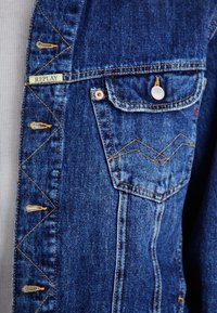 Denim jacket in dark blue with yellow stitching, featuring a front pocket and zigzag design along the button placket. Brand tag visible.
