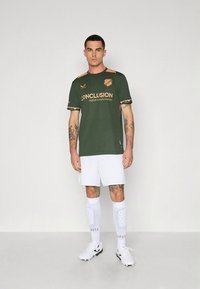 Grønn fotballtrøye med korte ermer og oransje tekst, hvite shorts og hvite knestrømper med prikkete tekstur. Atletenes sko er svarte.