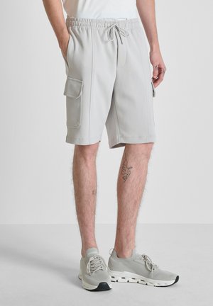 Hombre con pantalones cortos cargo de color gris claro con cintura ajustable con cordón y zapatillas grises a juego, de pie sobre un suelo gris.