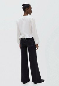 Top bianco a maniche lunghe con orlo arricciato e chiusura a chiave sul retro, abbinato a jeans neri a vita alta e gamba larga. Tessuto liscio con dettagli minimi.