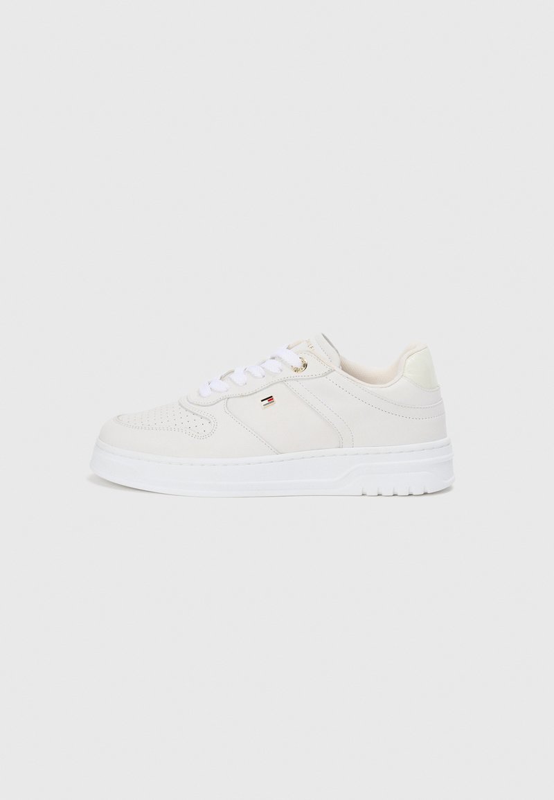 Baskets blanches montantes avec un lacage à l'avant, un bout perforé et un petit logo rouge, blanc et noir sur le côté, sur fond uni.