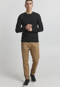 Zwarte longsleeve crewneck shirt met een textuur, gecombineerd met beige taps toelopende broeken en zwarte sneakers, staand op een lichte achtergrond.