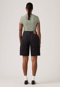 Γυναίκα με πλεγμένα μαλλιά που φοράει πράσινη και λευκή ριγέ μπλούζα, μαύρα τζιν σορτς, λευκές κάλτσες και μαύρα παπούτσια, στέκεται με την πλάτη γυρισμένη.