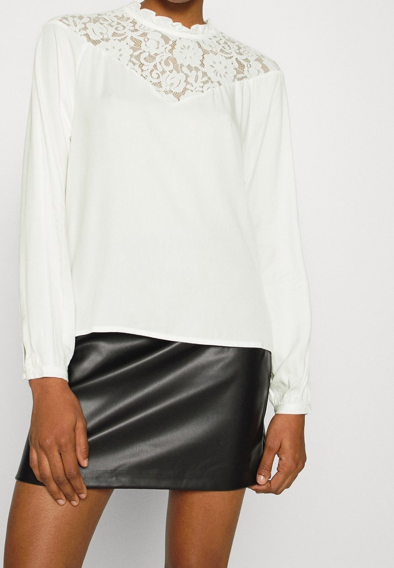 Femme portant un chemisier blanc à manches longues avec des détails en dentelle sur le haut et une mini-jupe en cuir noir, debout les mains près de l'ourlet de la jupe.