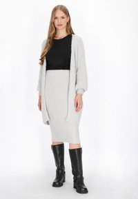 Lichtgrijze gebreide cardigan over een zwarte top en bijpassende geribbelde rok. Zwarte kniehoge laarzen met gespen maken de look compleet.