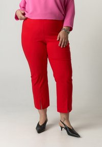 Femme portant un pantalon rouge court, un haut à manches longues rose, un bracelet en argent et des escarpins à talons recouverts noirs sur fond blanc.