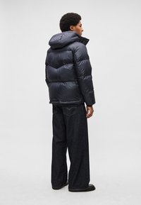 Casaco acolchoado azul-marinho com capuz, apresenta um design em espinha de peixe, textura brilhante e bainha canelada, combinado com jeans escuros de corte largo.