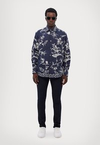 Camisa de botones en azul marino con estampado botánico blanco, combinada con jeans oscuros y zapatillas blancas. La camisa tiene cuello y mangas largas.