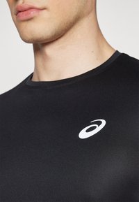 Svart sporttröja i slät framtill, med rund halsringning och en liten vit logotyp på bröstet. Närbild.