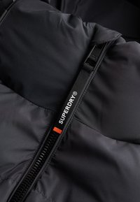 Černá péřová bunda s robustním zipem a výraznou štítkem s logem "SUPERDRY". Hladká, mírně lesklá tkanina s prošívanou texturou.
