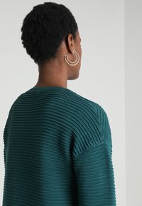 Teal ribbad tröja med texturerad design, rund halsringning och sänkta axelsömmar. Stora runda silverörhängen syns.