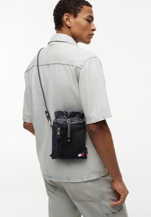 Mann trägt ein hellgraues Kurzarmhemd und eine Hose mit einer schwarzen Umhängetasche, die eine Reißverschlusstasche und das Tommy Hilfiger-Logo aufweist.