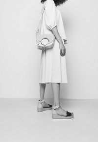 Sandales compensées beiges avec des franges noires, semelle en jute tissée, sangle de cheville assortie ; associées à une robe blanche fluide et un sac à épaule.