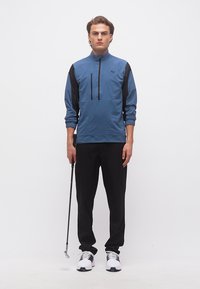 Άνδρας με αθλητικό μπουφάν σε μπλε και μαύρο χρώμα, μαύρο παντελόνι και λευκά αθλητικά παπούτσια κρατώντας ένα γκόλφ παύτερ, στέκεται μπροστά σε μια απλή λευκή επιφάνεια.