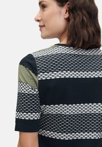 Chemise à manches courtes avec un motif composé de sections noires, blanches et vert olive ; tissu texturé avec des formes géométriques.