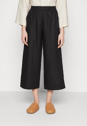 Personne portant un pantalon large noir coupé court, un haut beige à manches longues, et des chaussures tissées beige clair à enfiler, debout devant un fond blanc uni.