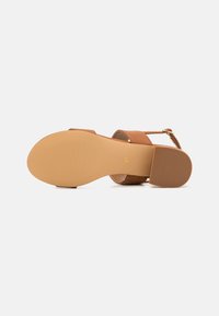 Sandal de cuero marrón con una correa texturizada y un tacón cuadrado de tamaño medio. La suela es de goma color tostado con un patrón estriado para mayor agarre.