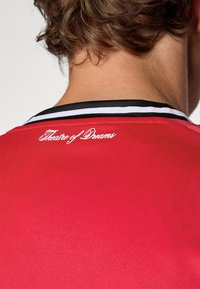 Piros sportmez fekete és fehér csíkos nyakkivágással, texturált anyagból, a „Theatre of Dreams” felirattal fehér hímzéssel.