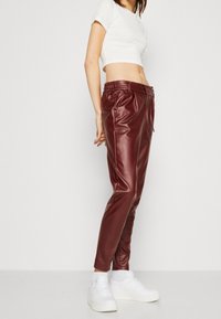 Pantalon en simili cuir bordeaux à coupe fuselée, taille élastique et détail cordon de serrage, porté avec des baskets blanches.
