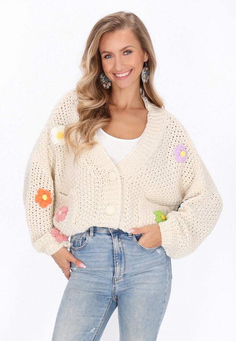 Cardigan lavorato a maglia di colore crema con appliqués floreali in arancione, rosa, viola e verde. Presenta un profondo scollo a V e due grandi bottoni.