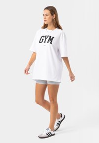 Bielo-čierna voľná tričko s nápisom "GYM" v tučných čiernych písmenách, kombinované so šedými športovými šortkami a klasickými čierno-bielymi teniskami.