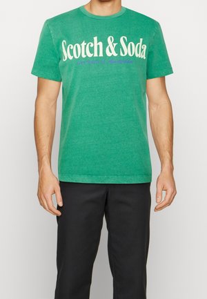 Homme portant un T-shirt à manches courtes vert avec "Scotch & Soda" imprimé en blanc et un texte plus petit en dessous, associé à un pantalon noir.
