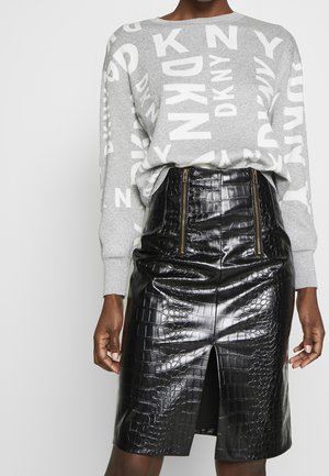 Personne portant un pull gris DKNY avec des lettres blanches et une jupe noire en simili cuir taille haute avec des zips à l'avant et une fente centrale.