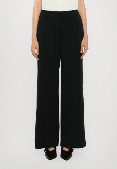 Pantalons noirs à jambes larges fabriqués dans un tissu lisse, avec une taille élastiquée à cordon de serrage et une coupe décontractée. Portés avec des chaussures noires à bouts pointus.