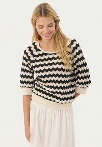 Pull rayé noir et crème avec un motif en vagues, col rond et manches trois-quarts, associé à une jupe blanche légère et fluide.