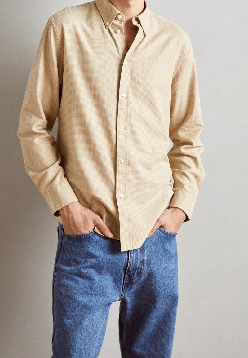 Homme portant une chemise beige à manches longues boutonnée avec col et un jean bleu, mains partiellement dans les poches, sur un fond clair uni.