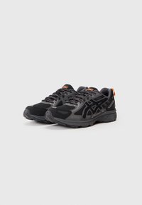 Um par de sapatilhas de corrida ASICS Gore-Tex em preto e cinza com acentos em laranja no logotipo, sobre um fundo leve e simples.