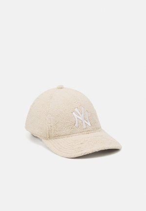 Beige fleece baseballpet met geborduurd wit "NY"-logo aan de voorkant en een gebogen klep tegen een effen lichte achtergrond.