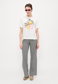 T-shirt bianca con grafica di limone e testo, abbinata a pantaloni larghi a righe bianche e nere e scarpe slip-on beige.